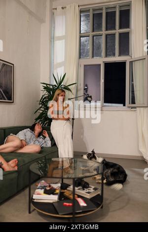 Drei Freunde genießen einen ruhigen Abend in einem gemütlichen Wohnzimmer, zwei ruhen auf einem Sofa aus und der andere lehnt sich auf den Arm des Sofas, als Nächstes mit einem verspielten Hund Stockfoto