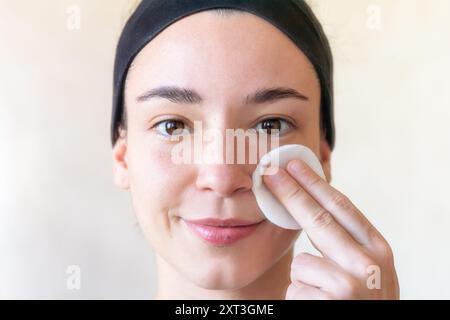 Eine junge Frau steht vor einem schlichten Hintergrund und wischt ihr Gesicht sanft mit einem weißen Wattepad ab. Sie trägt ein schwarzes Stirnband und hat eine natürliche, mak Stockfoto