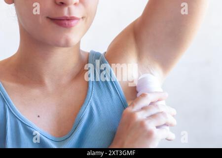Nahaufnahme einer Frau in einem blauen Tanktop, die ein Roll-on-Deodorant unter ihrem Arm aufträgt und so eine gesunde tägliche Selbstpflegeroutine unterstreicht Stockfoto