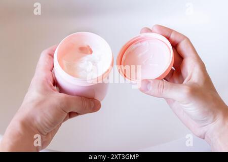 Hände halten ein Glas rosafarbener Feuchtigkeitscreme offen und konzentrieren sich auf ein Selbstpflegeritual in einer sauberen, hellen Umgebung Stockfoto