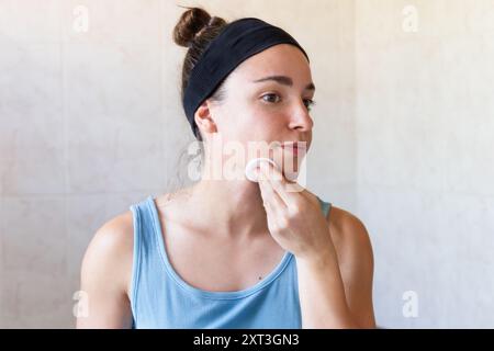 Eine ruhige Frau in lässigem Tanktop und Stirnband reinigt ihr Gesicht mit einem Wattepad in einem sonnendurchfluteten, hell gefliesten Badezimmer und verkörpert eine friedliche Atmosphäre Stockfoto