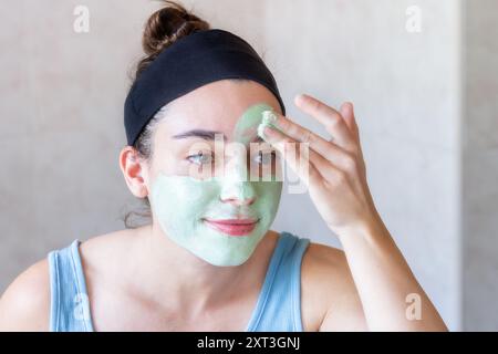 Eine junge Frau mit einem Stirnband trägt eine grüne Gesichtsmaske im Badezimmer und reflektiert einen Moment der Selbstpflege und Schönheitsbehandlung Stockfoto