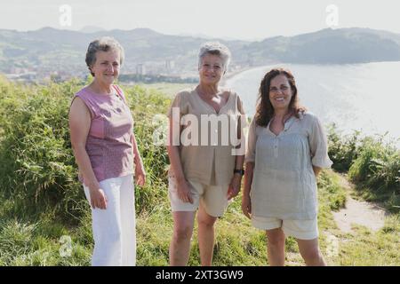 Drei ältere Frauen stehen zusammen im Freien mit einer Küstenlandschaft im Hintergrund. Alle lächeln in die Kamera, angezogen in legeren Sommer draußen Stockfoto