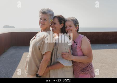 Drei ältere Freundinnen teilen sich eine warme Umarmung am Meer und fühlen sich fröhlich. Eine Frau schaut in die Kamera, während die anderen zwei nach unten schauen. Stockfoto