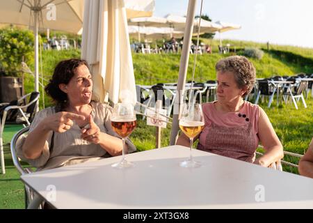 Zwei ältere, gehörlose Freundinnen führen ein lebhaftes Gespräch, während sie in einem Café im Freien ein Glas Bier halten, was beispielhaft für Gesellschaft und Entspannung ist Stockfoto