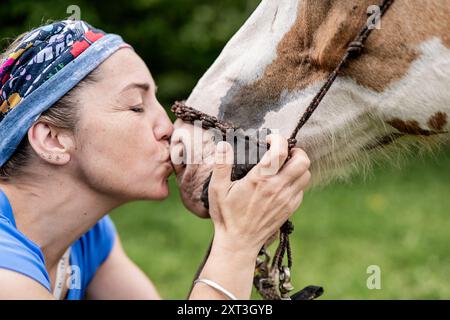 Nahaufnahme einer Frau, die liebevoll ein gemaltes Pferd auf den Maul küsst, stehend auf einer lebhaften grünen Weide. Das aktive, zärtliche Moment Captu Stockfoto