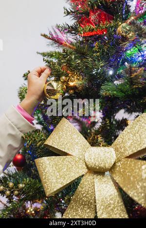 Ein kleines Kind reicht hoch, um ein goldenes Ornament an einen hell geschmückten Weihnachtsbaum zu hängen, der von leuchtenden Lichtern beleuchtet und mit einem großen Gold verziert ist Stockfoto