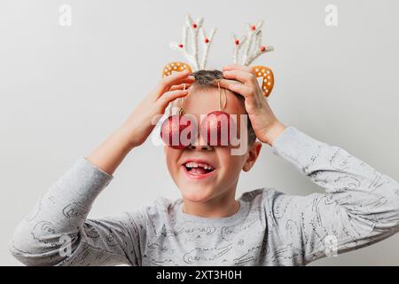 Ein fröhlicher kleiner Junge mit Rentiergeweih bedeckt seine Augen mit zwei rot leuchtenden Weihnachtskugeln vor neutralem Hintergrund Stockfoto