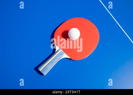 Ein lebendiges Bild, das ein rotes Tischtennispaddel mit einer weißen Kugel aufnimmt, hervorgehoben vor einem hellblauen Hintergrund. Die Komposition ist einfach und klar Stockfoto