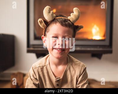 Ein fröhlicher kleiner Junge, der Rentiergeweih trägt, lächelt hell, sitzt zu Hause am glühenden Kamin und feiert den festlichen Geist des Weihnachtsstils Stockfoto