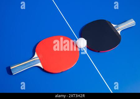 Dieses Bild zeigt zwei Tischtennispaddles, eines rot und eines schwarz, mit einer weißen Kugel dazwischen auf einem leuchtend blauen Tisch, die den Kontrast unterstreicht Stockfoto