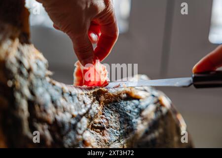 Eine detaillierte Nahaufnahme erfasst eine Hand, die ein Stück Serrano-Schinken aus einem gewürzten Bein sorgfältig schneidet, die Textur des Fleisches und die Präzision des Schnitts Stockfoto