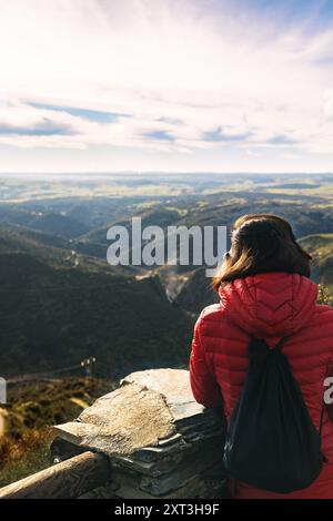 Eine Frau in roter Jacke steht am Aussichtspunkt Las Janas in Salamanca und blickt über eine weite Landschaft mit sanften Hügeln und üppigen Tälern, die im Sonnenlicht getaucht sind Stockfoto
