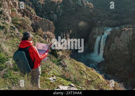 Eine Wanderer, die in einer roten Jacke gekleidet und mit einem Rucksack ausgestattet ist, steht mit einer Karte und blickt auf den majestätischen Wasserfall am Pozo de los Humos in Stockfoto