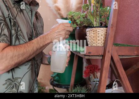 Ein Mann in einem tropischen Hemd tränkt Pflanzen mit einer Sprühflasche und konzentriert sich auf die Pflege einer gesunden Gartenumgebung Stockfoto