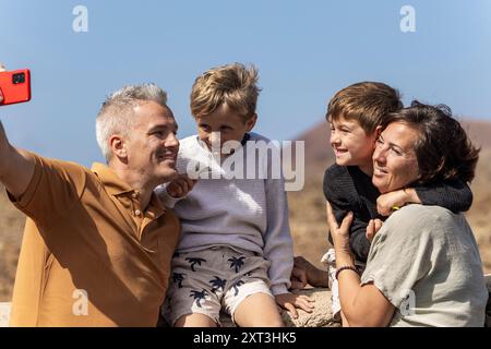 Eine vierköpfige Familie mit Mutter, Vater und ihren beiden Jungs genießt einen sonnigen Tag im Freien. Der Vater macht ein Selfie mit einem Smartphone, Captu Stockfoto