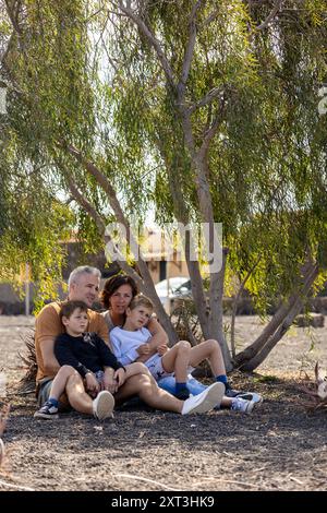 Eine vierköpfige Familie, Mutter, Vater und zwei Söhne entspannen sich im Schatten eines großen Baumes in einem Park Stockfoto