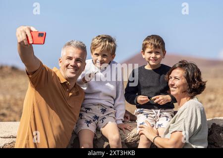 Eine vierköpfige Familie mit Mutter, Vater und ihren beiden Jungs genießt einen sonnigen Tag im Freien. Der Vater macht ein Selfie mit einem Smartphone, Captu Stockfoto