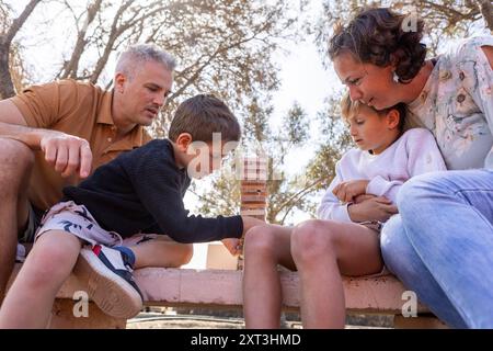 Von unten wird eine vierköpfige Familie mit einer Mutter, einem Vater und ihren beiden Söhnen gefangen, die sich bei einem Jenga-Spiel im Freien in einem Park befindet. Alle Studienteilnehmer sind c Stockfoto