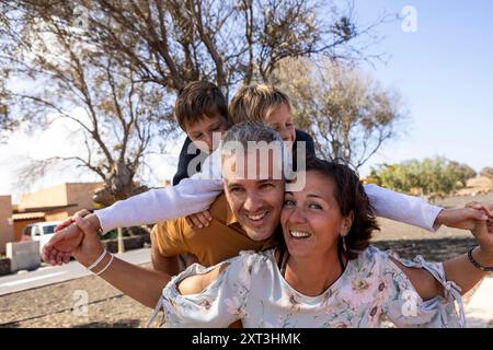 Glückliche Familienmomente, während Mutter und Vater mit ihren beiden Söhnen in einem Park spielen. Der Vater wird mit den beiden Söhnen auf dem Rücken gesehen. Stockfoto