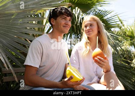 Ein junges Paar teilt sich einen freudigen Moment im Schatten üppiger Palmblätter und schlürft an einem sonnigen Tag erfrischende tropische Kokosnussgetränke Stockfoto