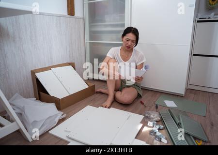 Eine junge asiatische Frau sitzt auf dem Boden und konzentriert sich auf Anweisungsblätter, während sie weiße Möbel zusammenbaut. Werkzeuge und Teile sind um sie herum verteilt Stockfoto