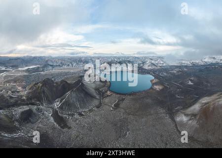 Atemberaubende Drohnenaufnahme eines ruhigen blauen Sees, eingebettet in das zerklüftete Gelände des isländischen Hochlands, mit dramatischen Landschaften unter einem bewölkten Himmel. Stockfoto