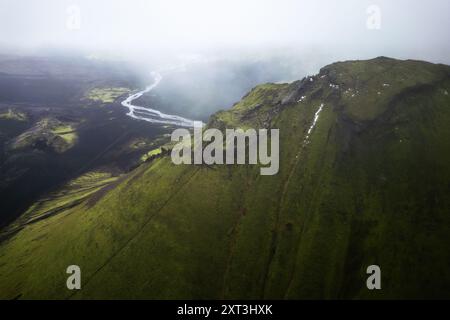 Eine Drohne fängt die atemberaubende Weite des Vulkans Mælifell in Island ein, mit nebeligem Himmel, einem mäandernden Fluss und lebhaftem Grün Stockfoto
