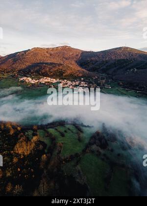 Ein Blick aus der Vogelperspektive fängt einen ruhigen Morgen mit Nebelschwaden über einem kleinen Bergdorf in Palencia ein, eingebettet zwischen den üppigen Gipfeln von Curavacas und Stockfoto