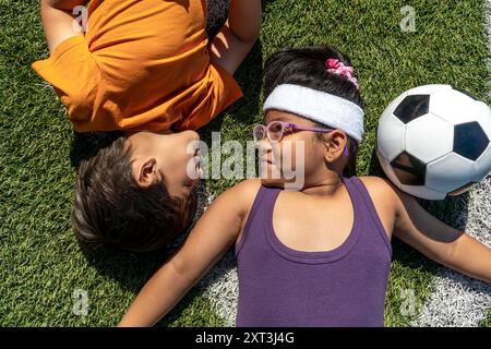 Zwei multiethnische Kinder, ein Junge und ein Mädchen, liegen auf grünem Gras neben einem Fußball, lächeln einander unter sonnigen Bedingungen an und stellen ein Spiel dar Stockfoto