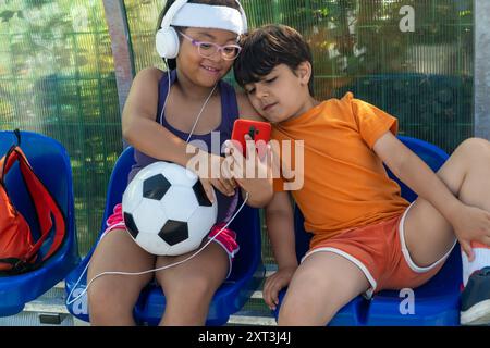 Zwei multiethnische Kinder sitzen eng auf blauen Stühlen und teilen sich ein Smartphone mit einem Fußball an ihrer Seite. Das Mädchen trägt Kopfhörer und einen lila ta Stockfoto