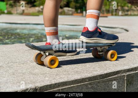 Nahaufnahme der Füße eines Kindes in Turnschuhen und Socken, die auf einem Skateboard mit gelben Rädern neben einem Brunnen stehen Stockfoto