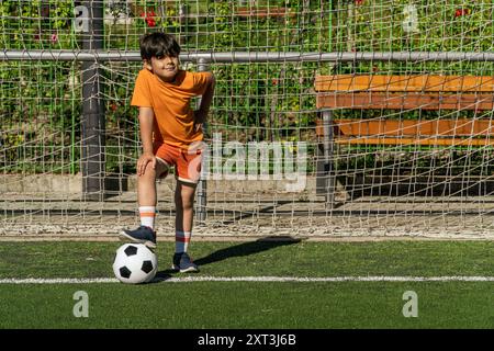 Ein kleiner Junge in einem orangefarbenen Hemd und Shorts steht an einem Fußballtor, der Fuß liegt auf einem Fußball und blickt selbstbewusst in die Kamera Stockfoto