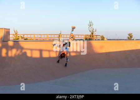 Ein junges Mädchen zeigt ihre Fähigkeiten im Rollschuhlaufen, indem es während des Sonnenuntergangs einen Handstand auf Rollschuhen in einem gut beleuchteten Skatepark macht Stockfoto