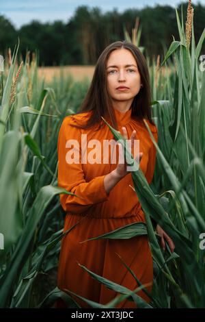 Eine brünette Frau in einem orangen Kleid steht nachdenklich inmitten großer grüner Maisstängel Stockfoto