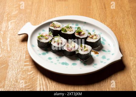 Eine künstlerische Präsentation von Sushi-Brötchen auf einer fischförmigen, weißen Keramikplatte, garniert mit grünen Sprossen und mit Sojasauce getränkt, wird auf gezeigt Stockfoto