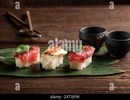 Ein Trio von Sushi-Stücken, elegant präsentiert auf einem breiten grünen Blatt, begleitet von dunklen Keramikschalen und Essstäbchen, die das Wesen des Japanischen einfangen Stockfoto