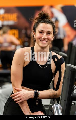 Eine junge Frau in Sportbekleidung macht eine Pause und lächelt sanft, während sie in einer lebhaften Umgebung in der Nähe der Geräte sitzt Stockfoto