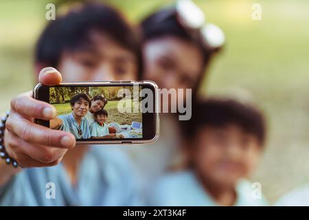 Eine asiatische Familie fängt einen fröhlichen Moment bei einem Picknick an einem sonnigen Sommertag ein. Der Vater hält ein Smartphone für ein Selfie in der Hand und zeigt ha Stockfoto