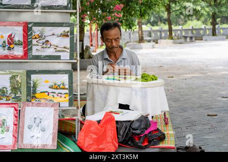 Ein erfahrener vietnamesischer Kanalisator arbeitet fleißig an detaillierten Stickereien, umgeben von farbenfrohen, gerahmten Kunstwerken in einer ruhigen Parklandschaft Stockfoto