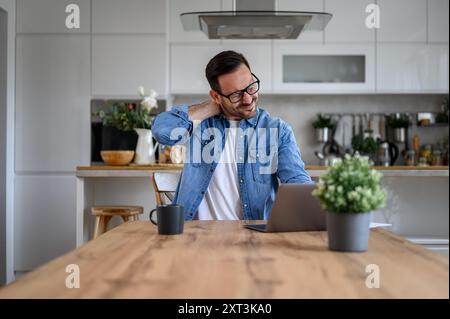 Erschöpfter junger Geschäftsmann, der den Hals vor Schmerzen berührt, während er am Schreibtisch im Heimbüro über einem Laptop arbeitet Stockfoto