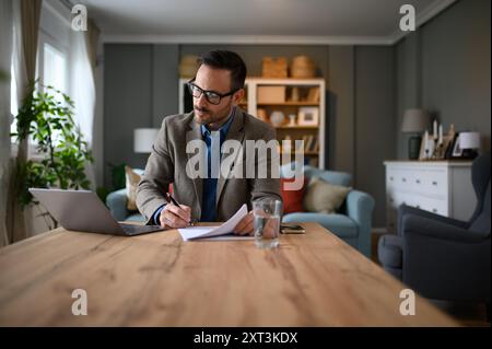 Fokussierter, eleganter Geschäftsmann, der Dokumente unterschreibt, während er im Heimbüro am Laptop arbeitet Stockfoto