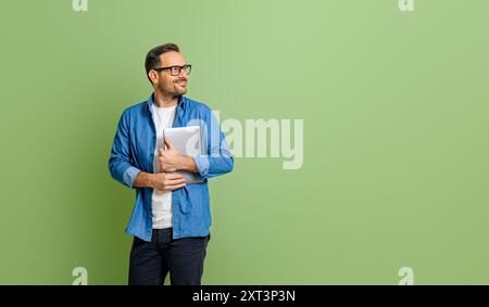 Junger, lächelnder Geschäftsmann, der ein digitales Tablet hält und nachdenkt, während er auf grünem Hintergrund wegschaut Stockfoto
