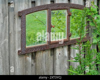 Rostiger schmiedeeiserner Fensterrahmen mit Spiegelglas an einer Schuppenwand, reflektierender Garten und Himmel. Ein Hauch von Nostalgie und vergangener Schönheit. Stockfoto