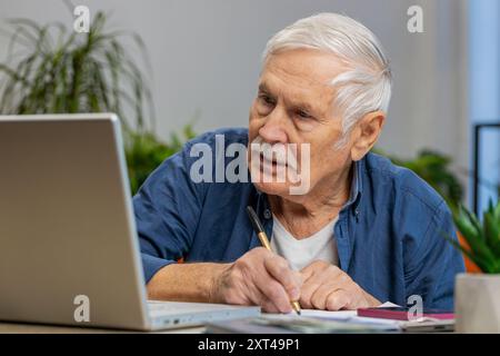 Senior man Buchhaltungszahlungen berechnen monatliche Ausgaben Bankrechnungen analysieren Papierkram in der Wohnung drinnen. Älterer, reifer Großvater, freiberuflicher Online-Job auf einem Laptop am Schreibtisch Stockfoto