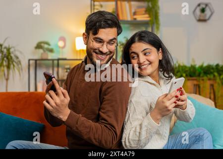Seitenansicht eines fröhlichen indischen Paares in legeren Kleidern, das sich hinten an hinten mit Smartphones auf dem Sofa im Wohnzimmer zu Hause sitzend. Junge Familie, die sich unterhalten, während sie ihr Handy auf der Couch in der Wohnung durchsucht. Stockfoto