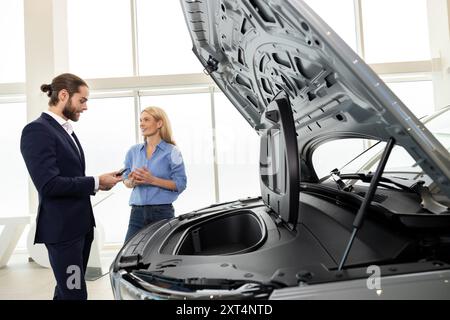 Autohändler beschreiben dem männlichen Käufer den Motor im Ausstellungsraum neben dem Fahrzeug mit geöffneter Motorhaube Stockfoto