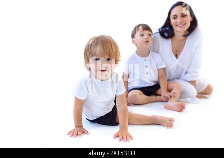 Eine junge schwangere Frau in einem weißen Kleid mit ihren süßen Söhnen. Die niedliche Mutter und ihre kleinen Kinder spielen auf dem Boden. Weißer isolierter Hintergrund. A ha Stockfoto