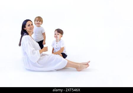 Eine junge schwangere Frau in einem weißen Kleid lächelt und sitzt mit ihren Söhnen auf dem Boden. Weißer isolierter Hintergrund. Lachende attraktive Mom und sie Stockfoto