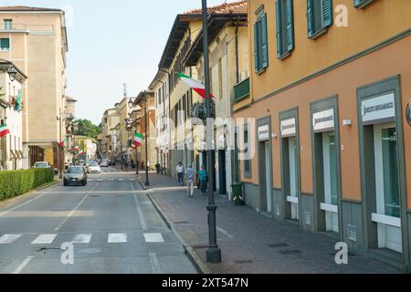 Rovato, Italien - 8. Juni 2023: Eine lebhafte Straße mit farbenfrohen Gebäuden und italienischen Flaggen, die an einem warmen, sonnigen Tag voller Fußgänger ist. Stockfoto